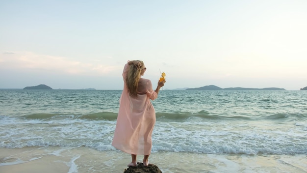 Attractive woman stand on a reef rock stone in sea on golden sunset Girl on tropical beach in green swimsuit and flutter in wind pink tunic silk shirt cape drinks her orange cocktail Pina Colada