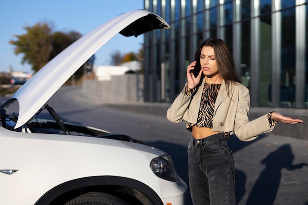 Attractive woman talking on the phone standing near a car with an open hood spreading her hands outdoors