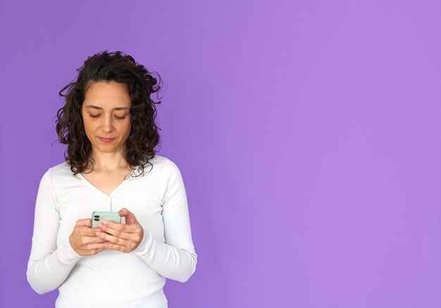 Attractive woman texting on her phone. Purple background and copy space. White casual shirt. Curly hair.