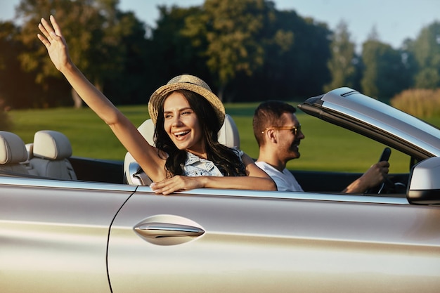 Attractive woman waving hand with man driving luxury cabriolet