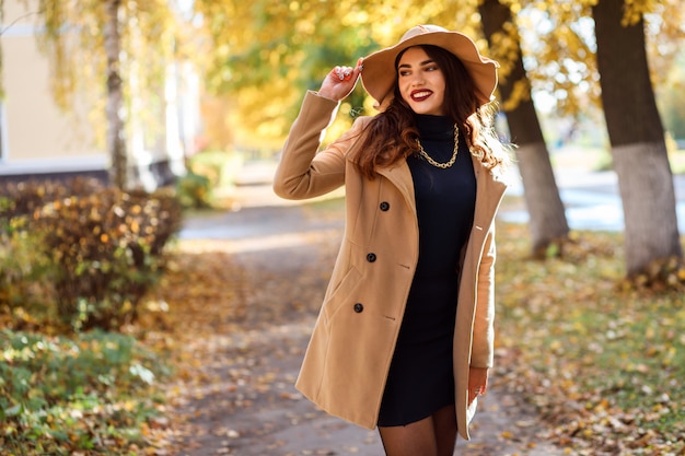 Photo attractive woman wearing wide-brimmed hat and beige coat is spending time outdoors in autumn. portrait of fashionable woman in fall season.