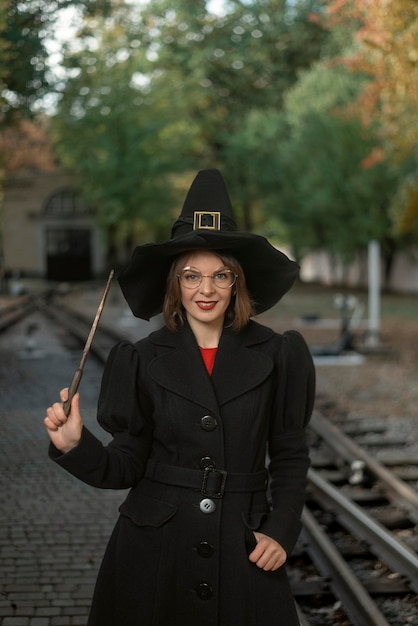 Attractive woman with wand in black pointed hat and coat on rails background Halloween celebration Vertical frame