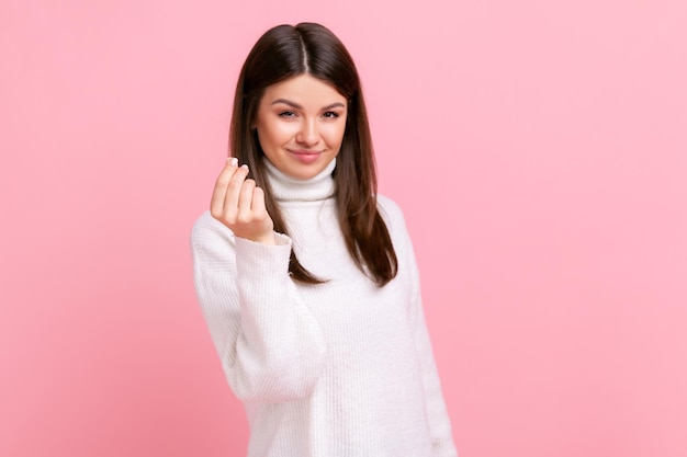 Attractive young adult woman standing and looking at camera with money or italian gesture with hand wearing white casual style sweater Indoor studio shot isolated on pink background