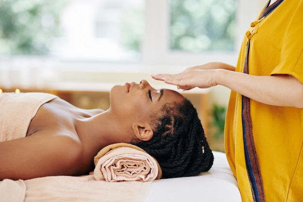 Attractive young African-American woman getting reiki healing therapy in spa salon