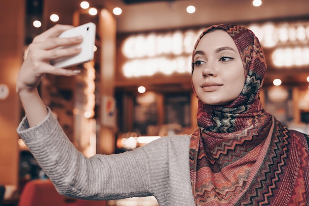 Photo attractive young arab woman with a beautiful headscarf sitting in an oriental restaurant and making selfie on her smartphone