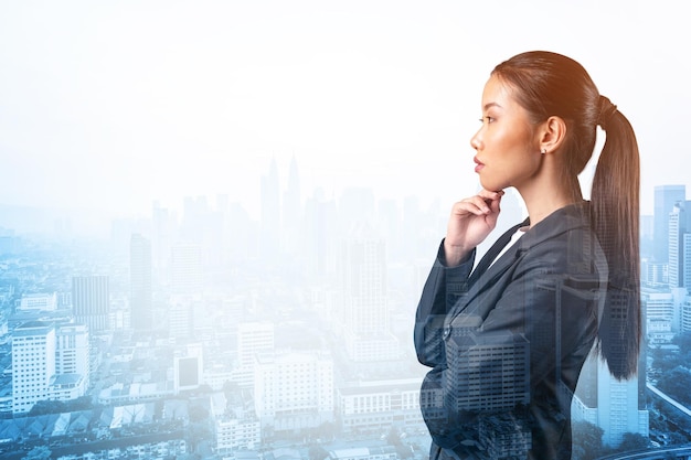 Attractive young Asian business woman in suit with hand on chin thinking how to succeed new career opportunities MBA Kuala Lumpur on background Double exposure