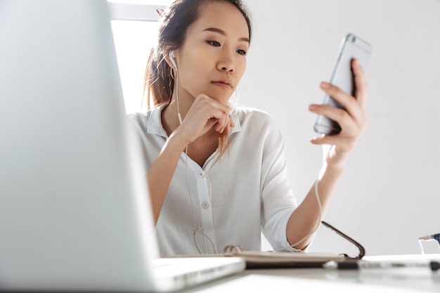 Attractive young asian businesswoman sitting at the office with laptop computer holding mobile phone, wearing earphones