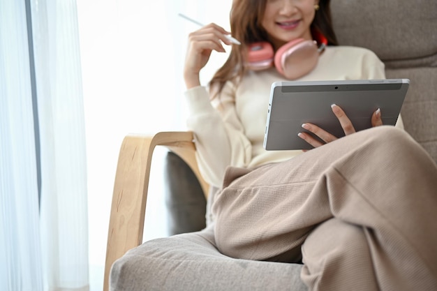 Attractive young Asian female with headphones relaxes on her comfy armchair while using tablet