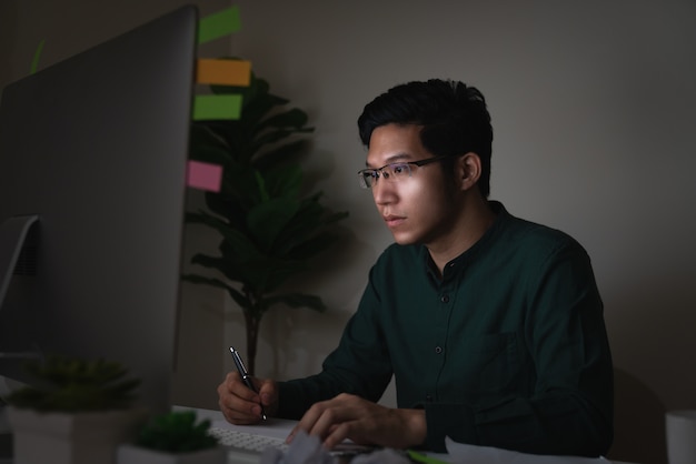 Attractive young asian man sitting on desk table looking at laptop computer in dark late night 