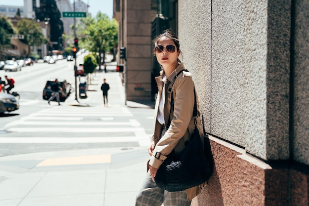 Attractive young asian woman in smart casual outfit standing on the road in san francisco under sunlight wearing sunglasses. chinese female against wall and looking into distance at her left side.