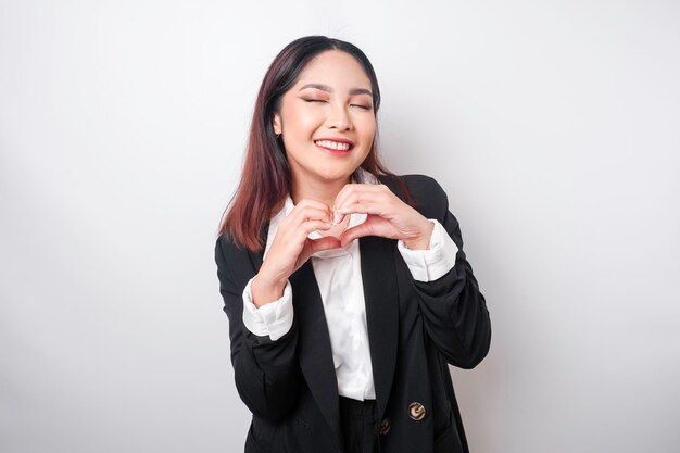 An attractive young Asian woman wearing a black suit feels happy and a romantic shapes heart gesture expresses tender feelings