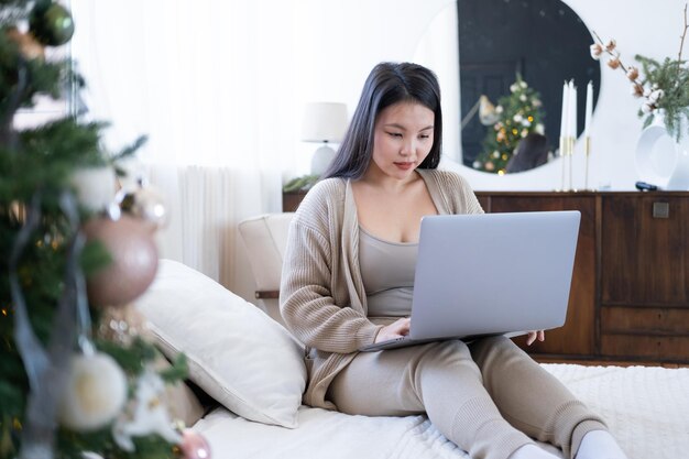 attractive young asian women in cozy clothing using laptop on bed at home, christmas time