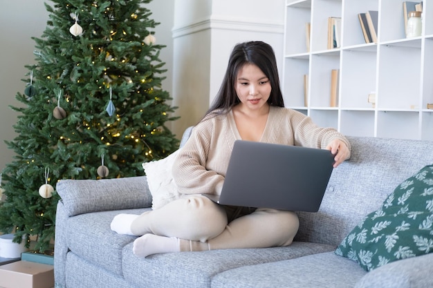 attractive young asian women in cozy clothing using laptop on couch at home, christmas time