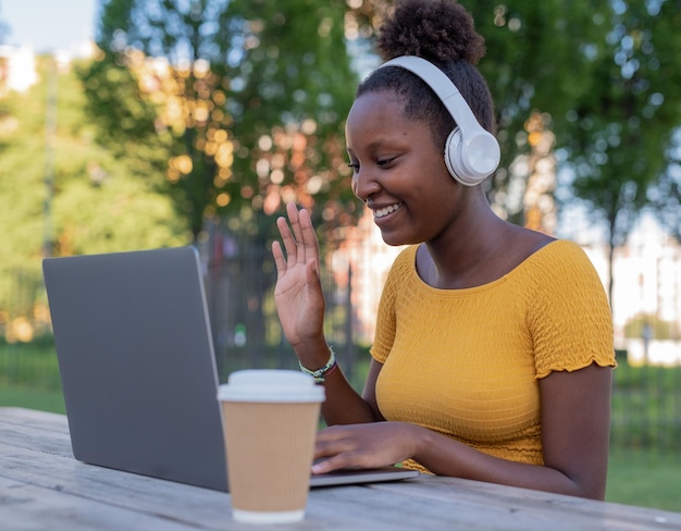 Attractive young black woman makes a video call in a park uses headphones greets friends distant relatives with her hands