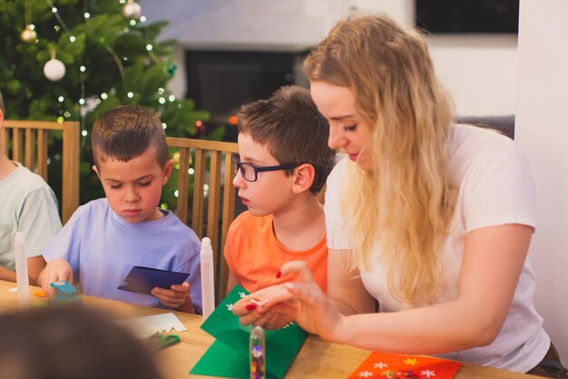 Attractive young blonde teacher helping pupils to cut from colorful paper Boys working concentrated with multicolor paper Christmas handmade workshop