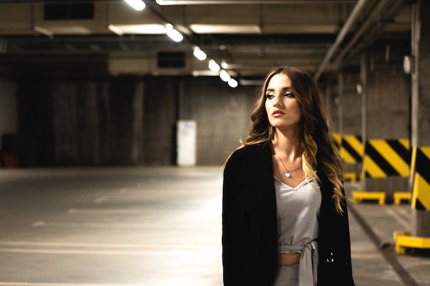 Attractive young brown-haired woman posing in an underground parking. Girl model in black coat posing in an empty underground parking. Fashionable photo of young beautiful girl with evening make-up