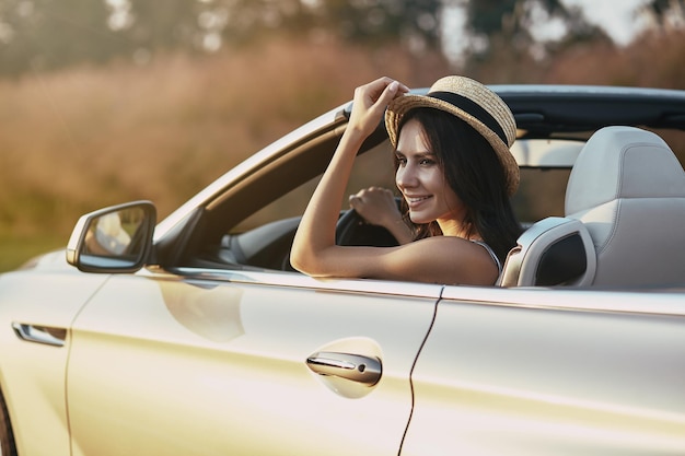 Attractive young brunette driving cabrio and holding hand on the hat looking away