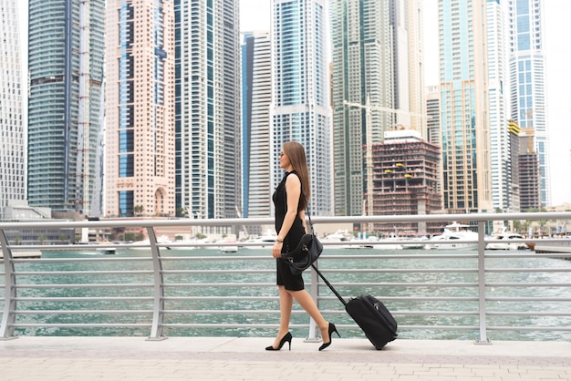 Attractive young business woman pulling suit case trough the magnificent city. Beautiful city scape panoramic view.