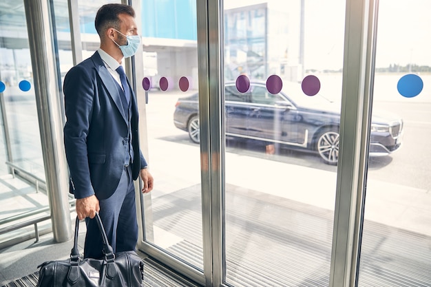 Attractive young businessman wearing a medical mask while standing with his luggage in the entrance hall of an airport and looking outdoors