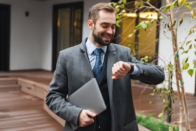 Attractive young businessman wearing suit walking outdoors, carrying laptop computer