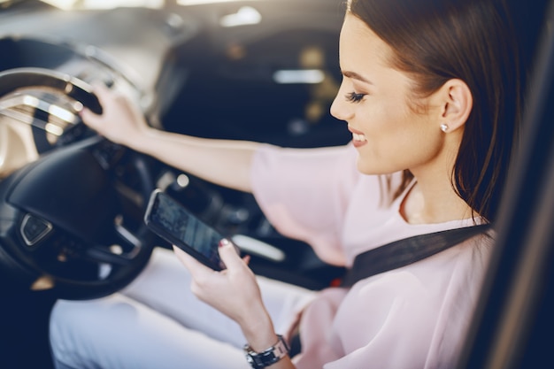 Attractive young Caucasian brunette with toothy smile and dressed elegant driving car and looking at smart phone.