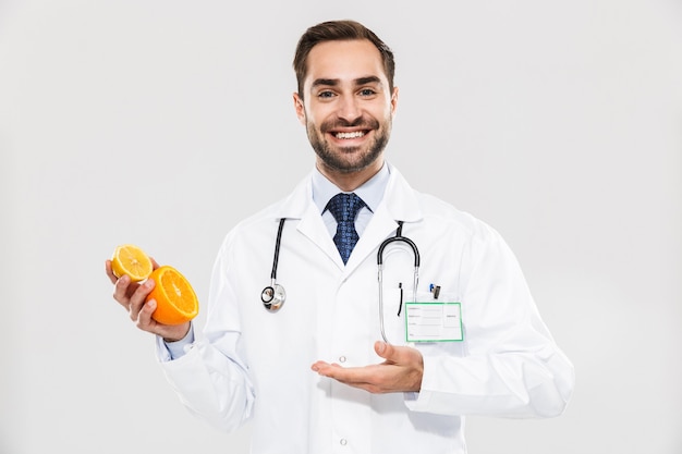 Attractive young cheerful male doctor wearing unifrom standing isolated over white wall, showing sliced orange