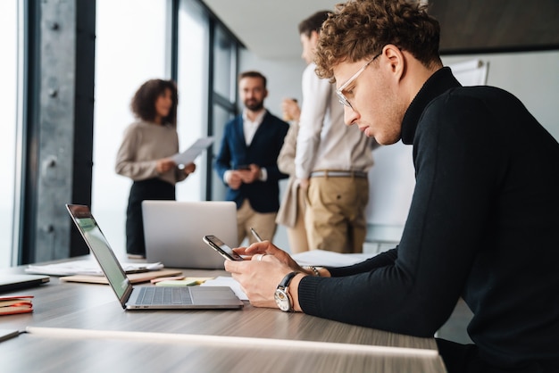 Attractive young confident businessman sitting at the office table with group of colleagues in the wall, using mobile phone