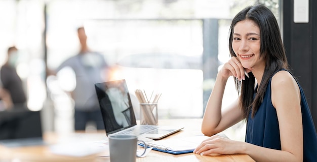 Attractive young confident designer sitting at the office table with group of colleagues in the background working on laptop computer