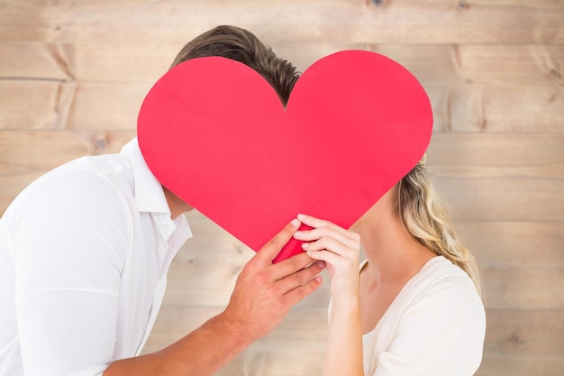 Attractive young couple kissing behind large heart against bleached wooden planks background