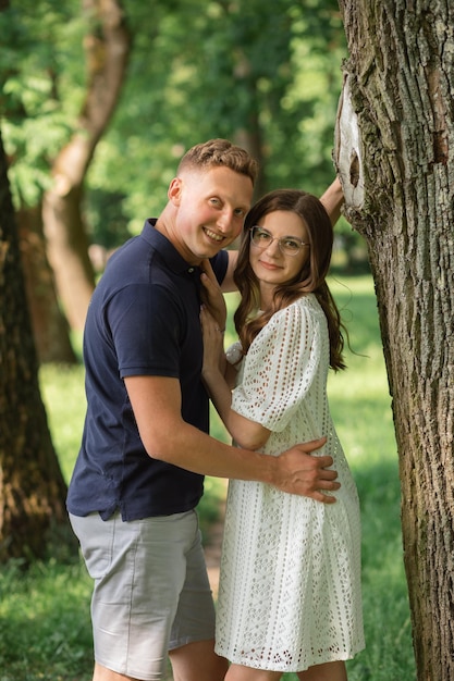 Attractive young couple in love embracing under trees in summer park looking at camera happy together