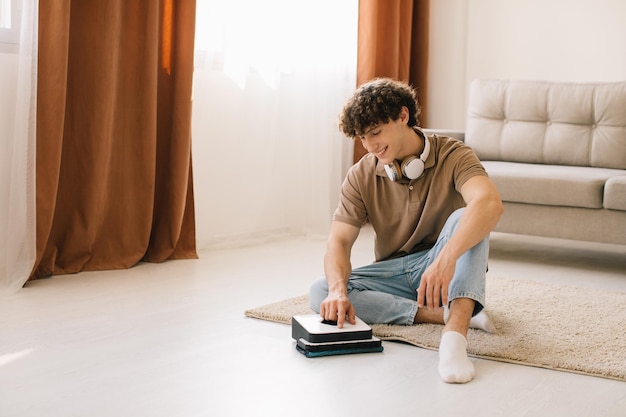 Attractive young curly man turning on washing cleaner robot while for cleaning carpet at living room at home