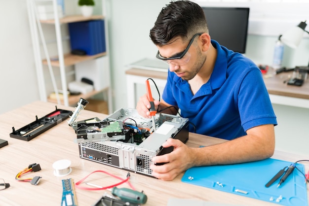 Attractive young engineer wearing protection glasses using a soldering iron to solder hardware on a computer at the repair shop