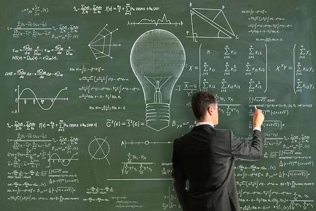 Attractive young european businessman drawing abstract lamp sketch with mathematical formulas on chalkboardblackboard wall background Intelligence idea solution science and innovation concept