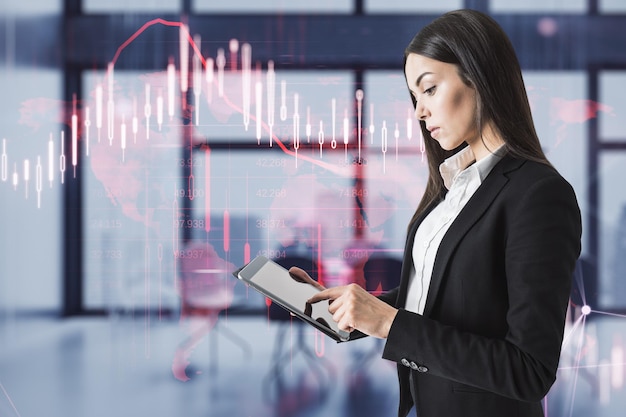 Attractive young european businesswoman using digital pad with glowing falling red forex chart on blurry office interior background Stock exchange and crisis concept Double exposure