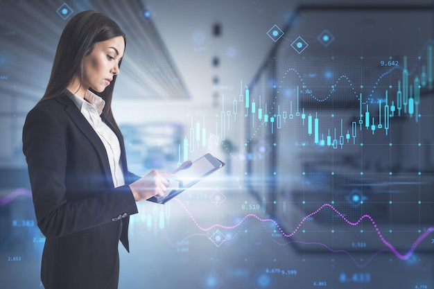 Attractive young european businesswoman using tablet while standing in blurry office interior with glowing forex chart hologram Trade finance and market concept Double exposure