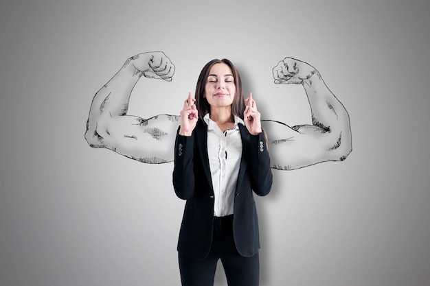 Attractive young european businesswoman with crossed fingers standing on concrete wall background with drawn muscle hands Strength leadership and independence concept