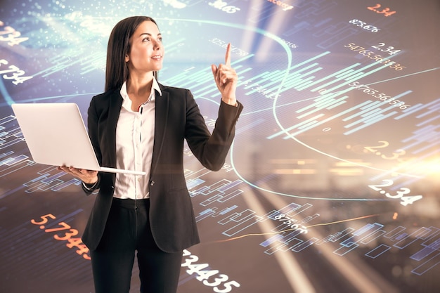 Attractive young european businesswoman with laptop pointing up on blurry office interior background with blurry forex chart Trade and market concept Double exposure