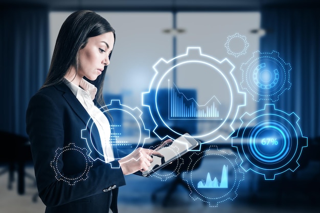 Attractive young european businesswoman with tablet and glowing cogwheel and chart hologram on blurry office interior background Business analytics and finance concept Double exposure