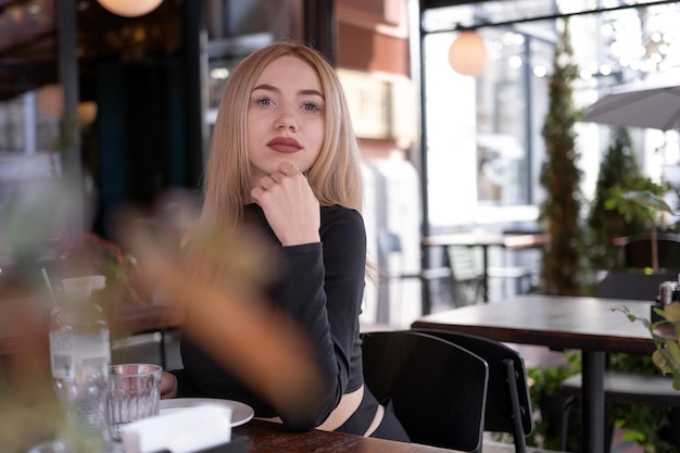 Attractive young fairhaired woman sits at table in cafe and looks pensively into camera Portrait of young girl in cafe