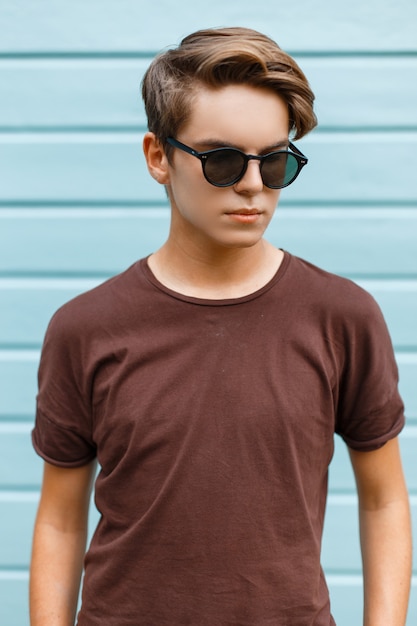 Attractive young fashionable hipster man in black stylish sunglasses in fashionable brown t-shirt poses in a city near a blue wooden building on a summer day