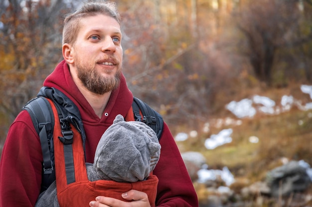 Attractive young father with his infant baby in sling fall outdoor Man is carrying her child and travel in nature