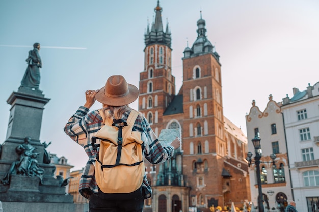 Attractive young female tourist is exploring new city Back view of girl in hat holding a paper map on Market Square in Krakow Traveling Europe at sunny day in autumn High quality photo