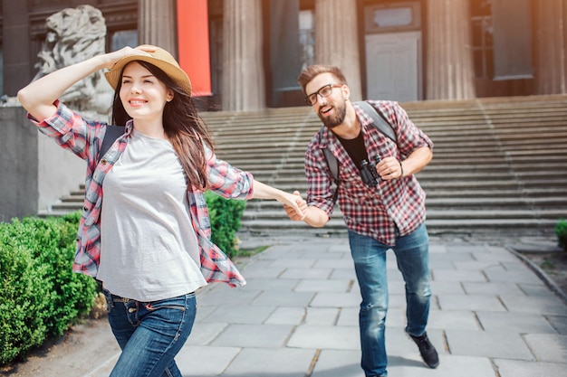 Attractive young female tourist walk forward. She smiles. Young woman hold boyfriend's hand and hat on here head at the same time.