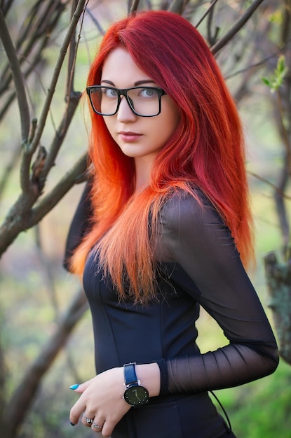 Attractive young girl with red hair wearing glasses posing near a tree