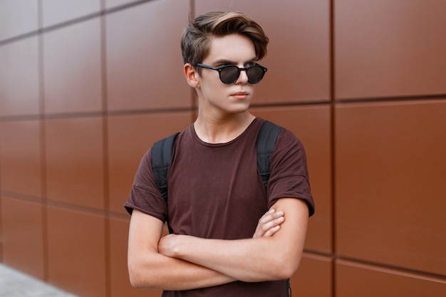 Attractive young hipster man in sunglasses in a summer t-shirt with a sport backpack is standing on a street on a sunny day near a vintage building