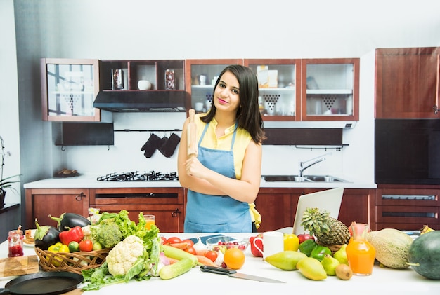 Attractive young Indian girl with apron cooking in kitchen with table full of fruits and vegetables