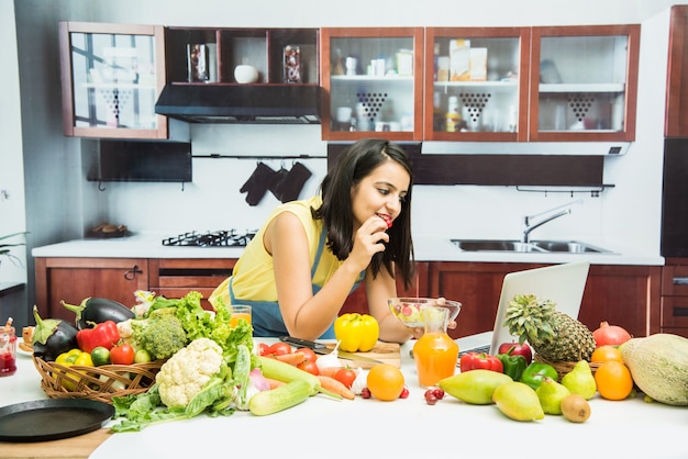 Attractive young Indian girl or woman with apron in kitchen multitasking, using smartphone, tablet pc with table full of fruits and vegetables and computer