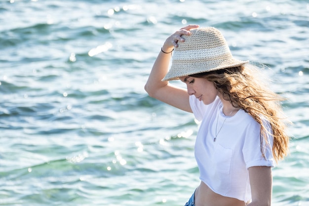 Attractive young latin woman portrait outdoors standing by the sea wearing a hat and summer clothes