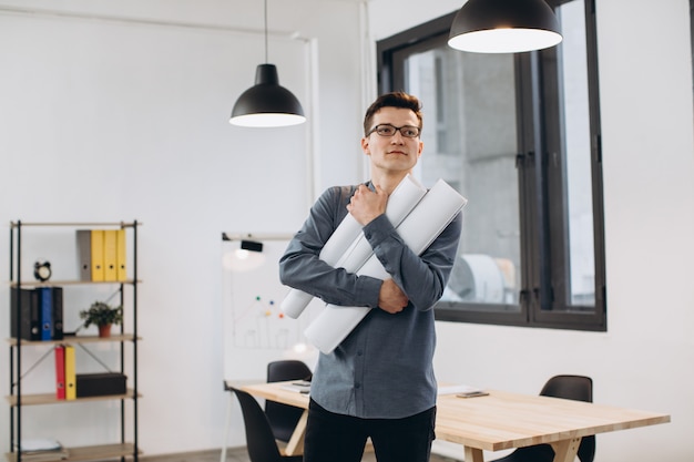 Attractive young man architect in glasses standing isolated over loft office, carrying drawings, holding house scheme