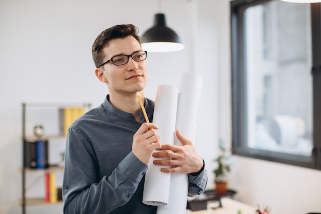 Attractive young man architect in glasses standing isolated over loft office, carrying drawings, holding house scheme